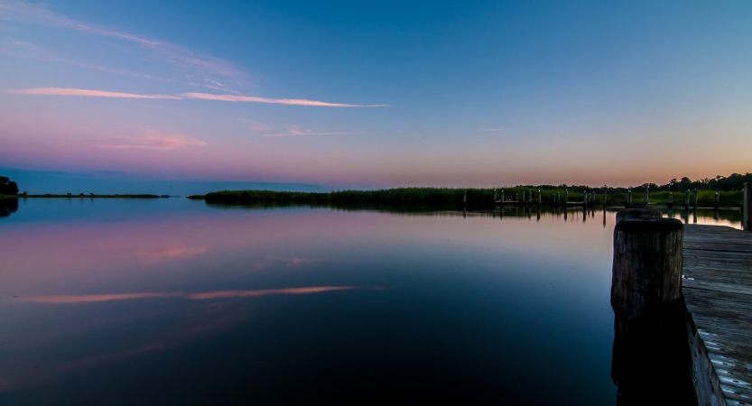 A very still body of water reflects the pink, purple and blue sky above. There is a dock in the foreground and trees lining the shore in the background. 